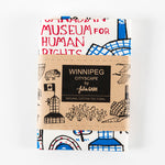 folded tea towel wrapped with a kraft brown paper band with the text “Winnipeg Cityscape by Julia Gash” 