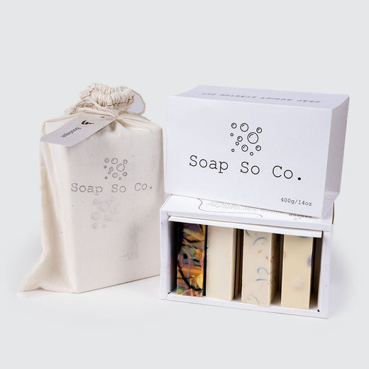 A white fabric bag with the brand name “Soap So Co.” written on it is on the left of an open box revealing four artisanal soaps in various colours. On top of the box is a white lid showcasing the brand’s name.