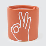 An orange-coloured cylindrical ceramic candle vessel features a white imprint depicting a hand making an “It’s OK” gesture with its thumb and index finger forming a circle and its middle, ring and pinky fingers extended.