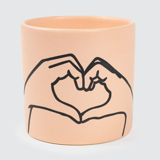 A coral-coloured cylindrical ceramic candle vessel features a black imprint of two hands forming a heart