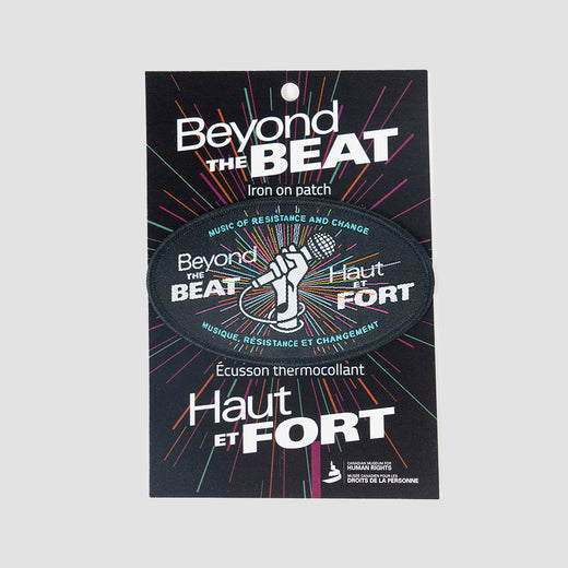 An embroidered black patch featuring a graphic of a hand holding a microphone and text inspired from the “Beyond the Beat” logo in English and French. It is shown on a black cardboard backing with similar graphics.