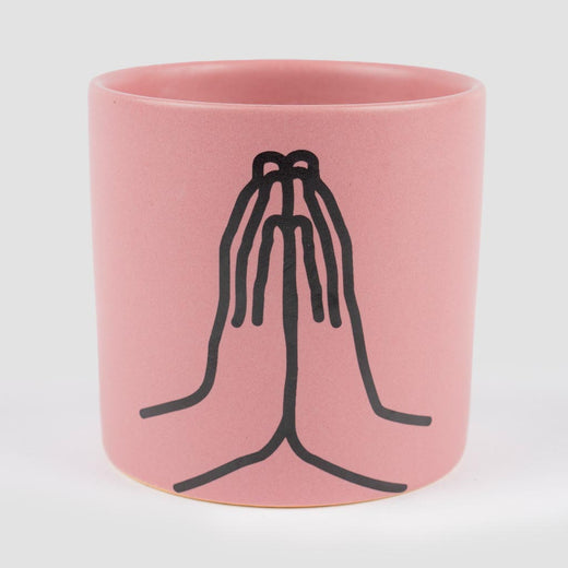 A pink-coloured cylindrical ceramic candle vessel features a black imprint of two hands forming a gratitude hand sign