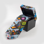 A tie in its box. The colourful print on the box top matches the print of the tie which is partially unrolled. 