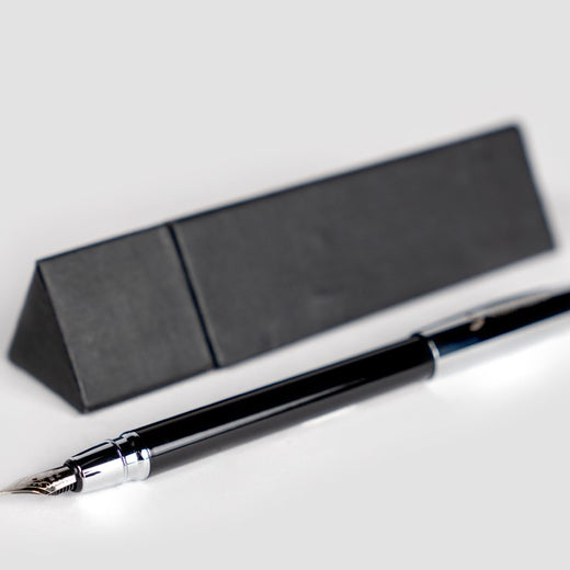 1. Black fountain pen lying on its side in front of its black triangle-shaped gift box. 2. Close-up of the pen’s chrome cap on which the Museum’s logo is engraved.