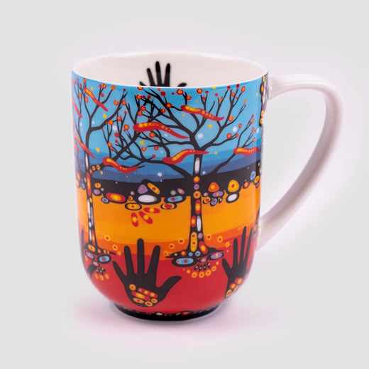 The front of the mug featuring the design. Hands painted in black circle the bottom third of the mug, with a red, orange, yellow, black, and blue gradient in the background. Trees painted in black are positioned in between each handprint.