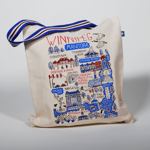 Front view of the canvas tote bag sitting upright. The blue, white, and red webbed handle is laying across the top of the bag to the left. The tote has a map of downtown Winnipeg in red, black, and blue.