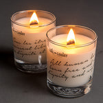 Two lit white candles with English and French writing etched on the glass containers. 