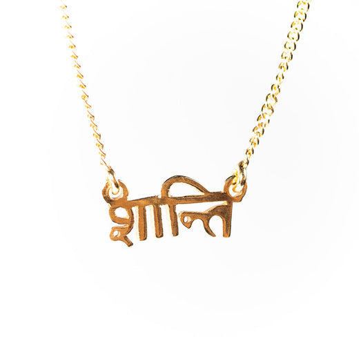 gold-coloured pendant on a chain; the pendant features the word “shanti” in Sanskrit