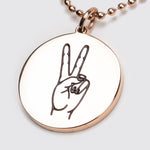 Rose gold circular pendant on which the hand sign for peace is engraved. It is suspended on a matching ball chain.