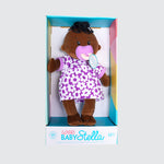 A dark-skinned cloth doll with a black ponytail on the top of its head. The doll is dressed in a purple and white dress with a matching soother in the mouth.